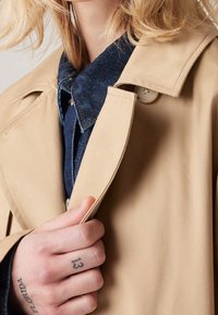 Beżowy trencz z zapinanym kołnierzem i klapami, noszony na ciemnoniebieskiej koszuli dżinsowej. Widoczna dłoń, tatuaż na palcu w pobliżu zapięcia. Miękka tekstura.