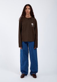 Braunes langärmliges Hemd mit einem Herz-Logo auf der Brust, kombiniert mit lässigen blauen Jeans. Glattes Material, entspannte Silhouette.