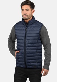 Marineblauw gewatteerd vest met hoge kraag en rits aan de voorkant. Grijze longsleeve shirt eronder. Soepele textuur met horizontale stiksels.