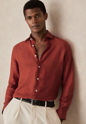 Jonge man die een open roestkleurig overhemd met knopen draagt, beige broek en een zwarte gevlochten riem, staand tegen een neutrale muur.