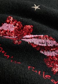 Tejido de punto negro con un lazo de lentejuelas en rojo y rosa, con texto bordado en rojo. La textura parece suave y esponjosa.