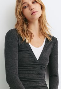 Grå langærmet top med V-hals designet lagdelt over en hvid T-shirt. Stoffet ser blødt ud med subtile ruching-detaljer ved taljen.