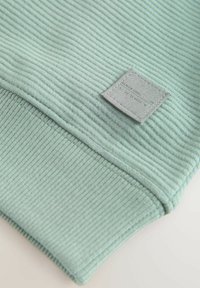 Tissu côtelé vert clair avec une texture douce, présentant une étiquette grise carrée cousue sur un poignet. La conception en vue de dessus met en valeur les détails du matériau.