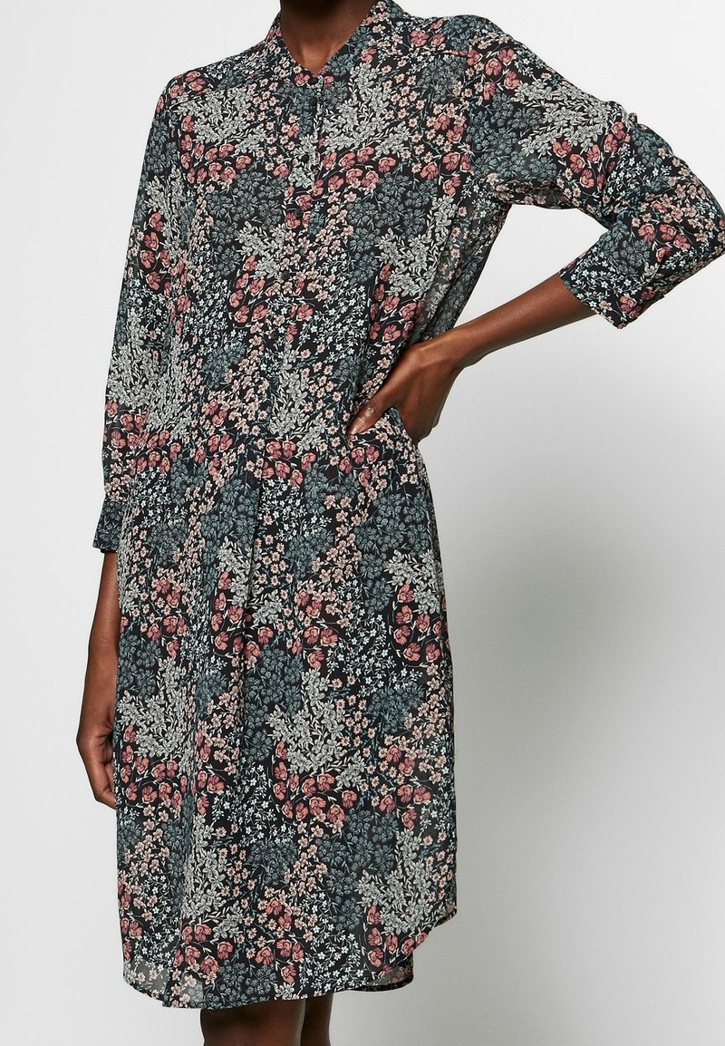 Robe noire à manches longues avec motif floral rose et blanc, col et boutons sur le devant, portée par une personne avec une main sur la hanche.