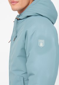 Lyseblå vandtæt jakke med hætte, der har et logo-patch på ærmet og snøreaccentuationer. Glat tekstur og moderne design.
