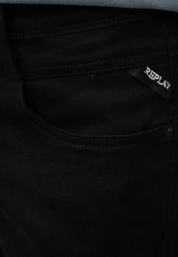 Τσέπη μαύρου τζιν με εμφανή ραφές και μικρή μαύρη ετικέτα με την επιγραφή "REPLAY" σε λευκό κείμενο.