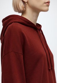 Personne aux cheveux blonds courts portant un sweat à capuche rouge foncé avec cordons, vue de profil du cou jusqu’au milieu du torse.