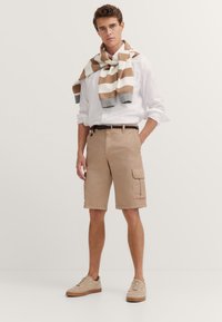 Beige cargo shorts with side pockets, paired with a white long-sleeve shirt and a striped brown and white sweatshirt draped over the shoulders.