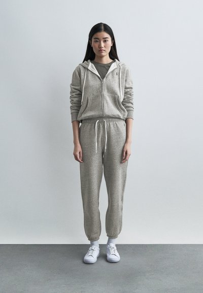 Νεαρή γυναίκα στέκεται μπροστά σε απλό τοίχο φορώντας γκρι φούτερ με φερμουάρ, ταιριαστά παντελόνια φόρμας, άσπρες κάλτσες και λευκά αθλητικά παπούτσια.