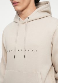 Mand iført beige hættetrøje med frontlomme og sort "JACK & JONES" tekst, vist fra bryst til hage i et nærbillede.