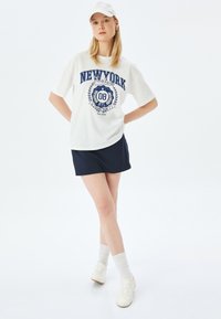 Hvid bomulds T-shirt med "NEW YORK" tekst i marineblå, parret med en marineblå nederdel, hvide sneakers og en hvid kasket. Enkel, afslappet design.