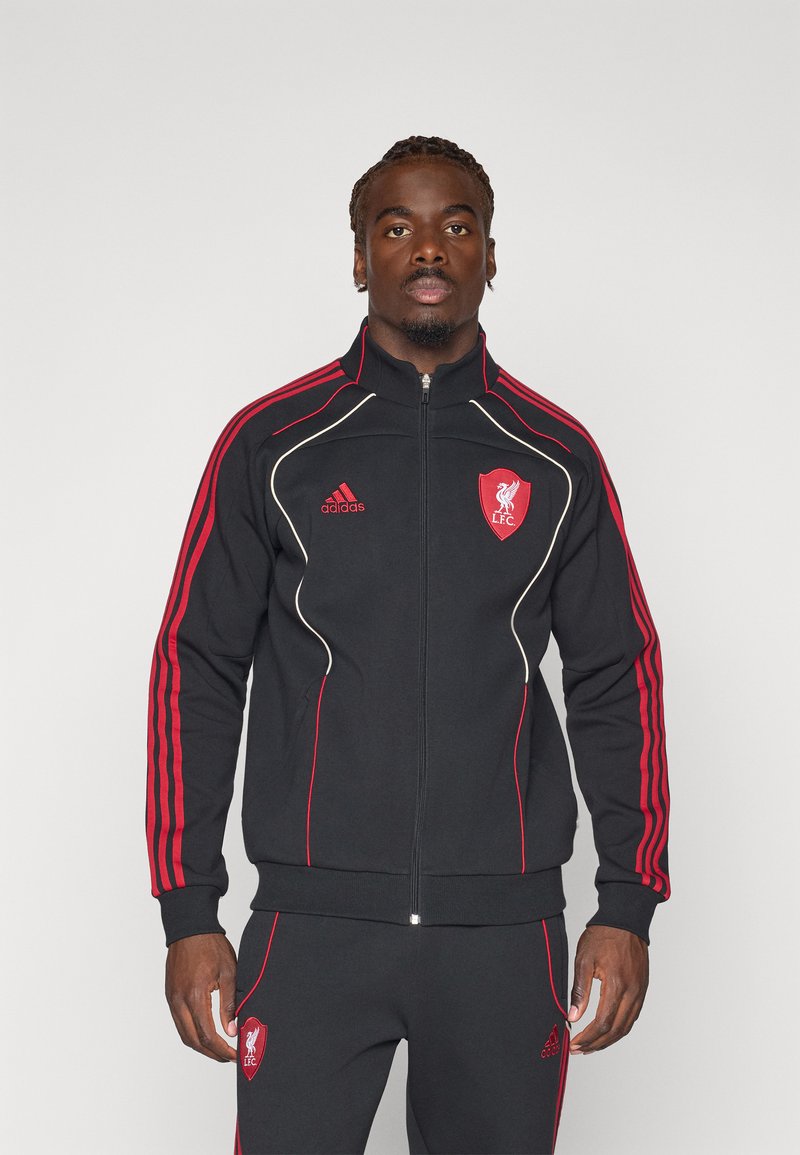 Melnas sporta uzvalks ar sarkaniem svītrām, ar rāvējslēdzēja aizdari un Liverpūles FC logo uz krūtīm; izgatavots no maiga auduma ar sportisku piegriezumu un ribotām aprocēm.
