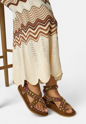 Sandales marron avec des clous colorés aux pieds, portant une longue jupe ondulée crème et marron, debout à côté d'une chaise en bois.