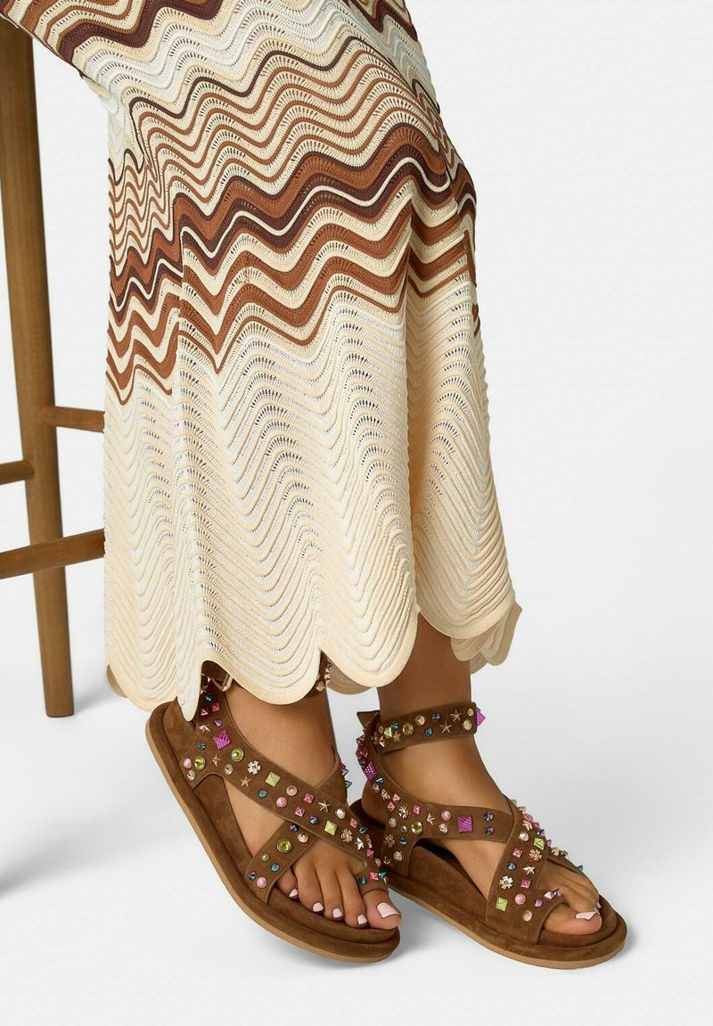 Sandales marron avec des clous colorés aux pieds, portant une longue jupe ondulée crème et marron, debout à côté d'une chaise en bois.