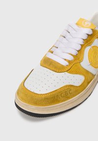 Tênis amarelo e branco com materiais de camurça e couro. Biqueira perfurada, cadarços brancos e sola de borracha preta texturizada. Emblema na lateral.