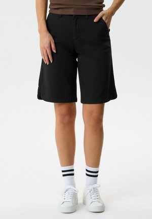 Personne portant un short noir jusqu'aux genoux, des chaussettes blanches avec des rayures noires, et des baskets blanches, debout avec une main dans la poche.