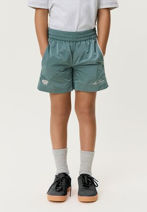 Kind trägt grüne Sportshorts mit gestickten Logos, graue Socken, schwarze Sneakers mit hellbraunen Sohlen, steht mit Händen in den Taschen.