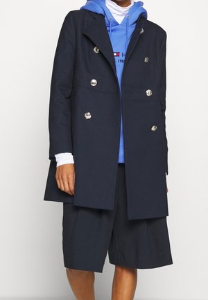 Person trägt marineblauen, zweireihigen Mantel über blauem Kapuzenpullover, weißem Langarmshirt und marineblauen, knielangen weiten Shorts vor schlichten Hintergrund.