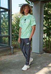 Camisa atlética de un verde claro con un patrón y cuello redondo, combinada con pantalones ajustados gris oscuro que tienen detalles y un logo en blanco. Zapatillas deportivas.
