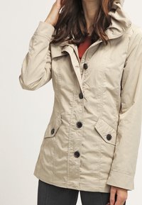 Chaqueta con capucha beige que presenta un cierre de botones en la parte delantera, dos bolsillos laterales y una tela ligeramente texturizada. Diseñada con un corte ajustado.