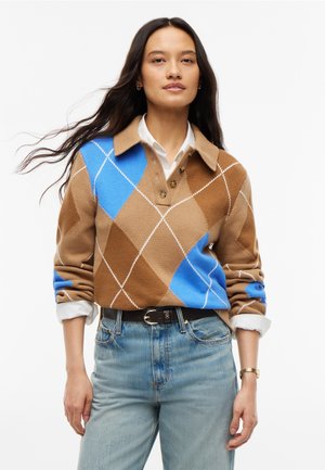Femme aux longs cheveux foncés portant un pull à losanges marron et bleu sur une chemise blanche, associé à un jean bleu clair et une ceinture noire.