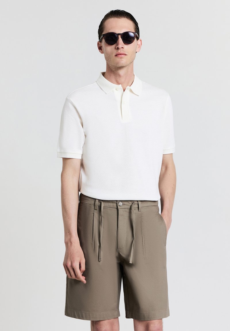 Weißes Poloshirt mit kurzen Ärmeln aus strukturiertem Stoff und einer drei-knöpfigen Knopfleiste, kombiniert mit khakifarbenen, gefalteten Shorts mit einem Gürtel und seitlichen Taschen.