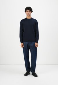 Jaunas vyras stovi priešais baltą sieną, apsirengęs tamsiai mėlynu megztiniu, mėlynomis džinsinėmis kelnėmis ir juodais odiniais batais, žiūrėdamas tiesiai, su neutraliu veido išraiška.