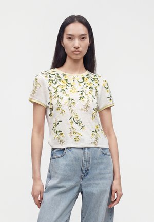 Femme aux cheveux noirs et raides portant un haut blanc à manches courtes avec imprimé floral jaune et un jean bleu clair taille haute.