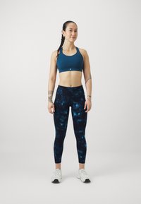 Mėlynos spalvos sportinis liemenukas, tamsiai mėlynos spalvos aukštai suknelės kelnės su galaktikos raštu ir baltos sportinės bateliai. Modelis stovi su atsipalaidavusiomis rankomis.