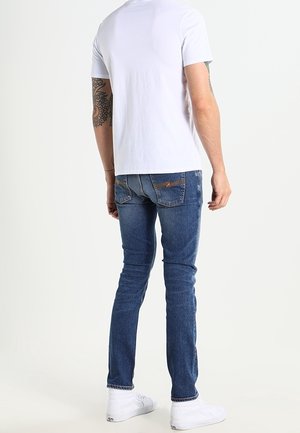 T-shirt blanc en coton associé à un jean en denim bleu, avec une coupe slim, des accents délavés et des baskets blanches. Vue de dos, fond neutre.
