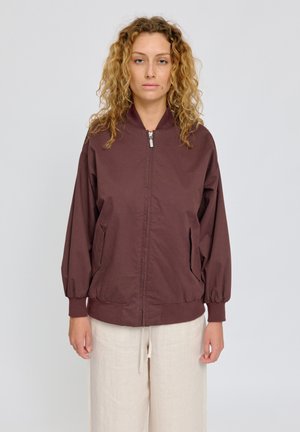 Woman with curly hair wearing a brown zip-up jacket and cream-colored pants standing against a plain light background.