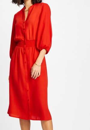 Femme portant une robe mi-longue rouge vif boutonnée avec des manches bouffantes et une taille froncée, debout devant un fond blanc uni.