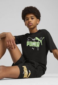 Camiseta negra con logo de Puma en verde y blanco en el frente, combinada con pantalones cortos negros que presentan texto de Puma en amarillo. Material de algodón suave.