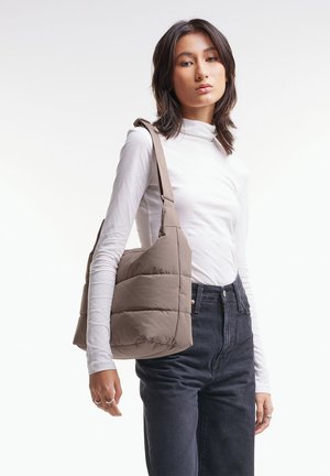 Frau trägt ein weißes langärmeliges Oberteil und schwarze Jeans, mit einer großen gepolsterten Schultertasche in Taupe auf ihrer rechten Schulter.