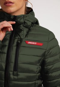 Zelená nadýchaná bunda s horizontálním prošíváním, černými zipy a červenou štítkem s logem "JACK1T" na levé straně hrudi. Hladký povrch.