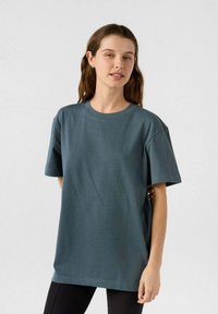 T-shirt in cotone verde scuro con maniche corte, scollo a giro e vestibilità ampia. Texture liscia senza motivi o dettagli visibili.