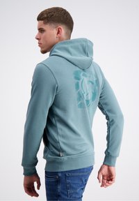 Zilganas džemperis ar kangaroo kabatu un ribotiem piedurknēm, ar lielu grafisku dizainu aizmugurē un ar auklu kapuci.