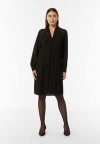 Robe noire plissée avec des manches longues, décolleté en V et lien en tissu. Texture lisse avec une silhouette fluide, portée avec des collants noirs et des talons.