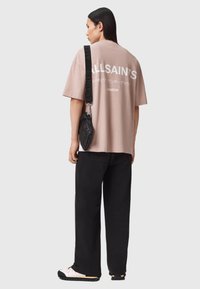 Hellrosa übergroßes T-Shirt mit fettem "ALLSAINTS"-Logo auf der Rückseite. Kombiniert mit schwarzen, locker sitzenden Hosen und einer kleinen schwarzen Umhängetasche.