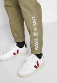 Zielono-oliwkowe dresy z elastycznymi mankietami i białym haftowanym napisem "KANI" po boku. W połączeniu z białymi sneakerami z czerwonymi akcentami logo.