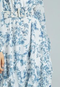 Blumenkleid in Weiß mit blauen Mustern, ausgestattet mit einem gerafften Saum und einem passenden Gürtel. Leichter Stoff mit weicher Textur.