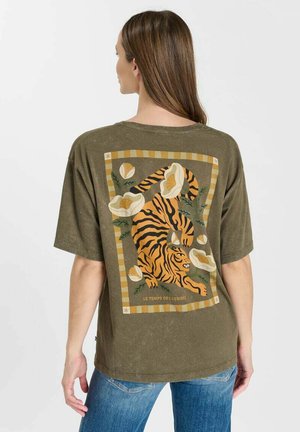 Femme portant un t-shirt vert olive avec un graphisme d'un tigre orange et noir entouré de fleurs blanches dans le dos.
