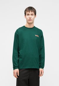 Młody mężczyzna z krótkimi ciemnymi włosami, ubrany w zieloną koszulę z długim rękawem z czerwonym tekstem graficznym na piersi, stojący na białym tle.