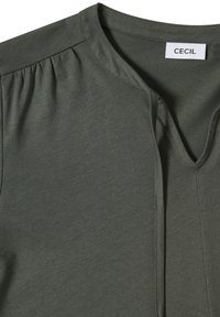 Haut à manches courtes vert olive avec un col en V, présentant une texture douce et lisse ainsi qu'un léger froncé aux épaules. L'étiquette indique "CECIL".