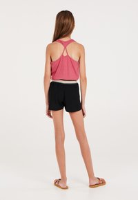 Top senza maniche rosso con design incrociato sulla schiena, abbinato a pantaloni shorts neri con vita elasticizzata e dettagli verdi. Sandali marroni.