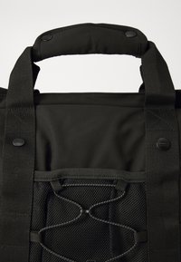 Črna platnena torba z oblazinjeno ročico, mrežasto žepom in elastiko za pritrditev predmetov. Teksturno zaključena s šivanimi detajli.