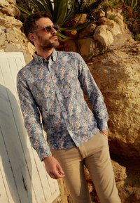 Een knoopshirt met een blauw bloemenpatroon en roze accenten. Gestyled met kaki broek en zonnebril tegen een rotsachtige achtergrond.