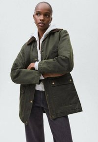 Oliivinvihreä takki ruskealla kauluksella, jossa on vetoketjusuljenta, napit ja kaksi suurta etutaskua. Päällä harmaa huppari.