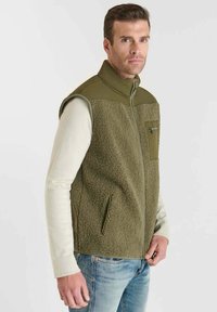 Olivgrüne Weste mit einer weichen, strukturierten Vorderseite, glattem Rücken, Reißverschluss und einer Brusttasche. Über einem hellen Langarmshirt getragen.