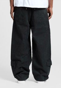 Persona che indossa pantaloni cargo neri larghi con tasche laterali, mostrata di spalle, in piedi su uno sfondo bianco semplice.